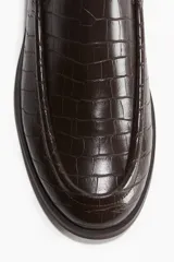 Mocasín de vestir color marrón oscuro con textura de piel de cocodrilo en relieve. Presenta un diseño clásico con costura tradicional en la pala y suela baja de goma.