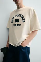 Sudadera boxy fit de color beige con interior de rizo, cuello redondo y manga corta. Presenta un estampado contrastante en el frente con la inscripción "Deep Founders London 90" y acabados irregulares en los bordes.