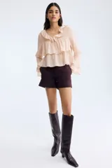 Blusa rosada de tejido liviano, con escote en V rodeado de volados y tiras finas para anudar. Presenta un diseño escalonado con volados anchos en el pecho y en el bajo, y mangas largas acampanadas con volados dobles en los puños.