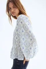 Blusa en mezcla de viscosa con peplum acampanado, cuello redondo, escote en V y tiras finas con cuentas decorativas. Presenta mangas globo largas con elástico en los puños y un estampado en tonos blancos y azules con motivos florales y geométricos.
