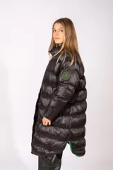 Campera puffer larga de color negro con acabado brillante, cierre frontal con botones a presión y cuello alto. Presenta un parche circular en la manga izquierda.