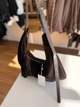 Cartera tipo hobo de textura gamuzada en color marrón oscuro, con diseño flexible y asa larga regulable. Presenta un detalle de flecos largos desmontables en uno de los laterales y cierre superior.