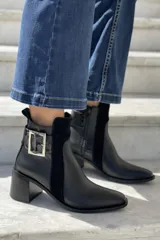 Botín de cuero color topo o gris-marrón, con punta afilada y taco ancho y alto de madera oscura. Presenta un diseño con tira y hebilla metálica cuadrada en el lateral exterior y cierre de cremallera en el lateral interior.