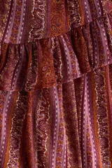 Vestido corto de manga larga confeccionado en tejido transparente, con estampado estilo paisley en tonos borravino y rosado. Presenta cuello redondo con abertura frontal y cordones para atar, cintura elástica y falda con volados en capas.