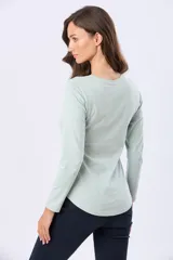 Remera de manga larga en color verde menta, con cuello redondo y cierre frontal parcial de botones a tono. Presenta un diseño con ruedo curvo y un pequeño aplique metálico en la parte inferior.