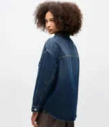 Camisa de jean estilo overshirt con corte oversized, cuello clásico y mangas largas. Presenta cierre frontal con botones, dos bolsillos delanteros con solapa y aplicación de tachuelas metálicas en el cuello y la parte superior del pecho.
