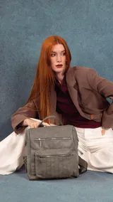 Mochila urbana de tamaño mediano confeccionada en cuero sintético color gris. Presenta un diseño con dos compartimentos principales con cierre, un bolsillo frontal con detalle de costuras verticales, asa superior y correas ajustables.