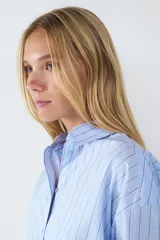 Camisa celeste con finas rayas verticales negras, de corte amplio, manga larga, cuello clásico, cierre frontal con botones y puños abotonados.