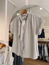 Blusa de rayón color gris claro, con escote en V, cierre frontal con botones y un lazo integrado en el cuello. Cuenta con mangas cortas con vuelta y diseño fluido.