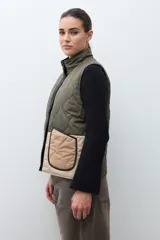Chaleco capitoneado reversible con cuello alto, cierre frontal con botones a presión y bolsillos delanteros. Presenta un diseño bicolor en tonos verde oliva y beige con detalles en negro.