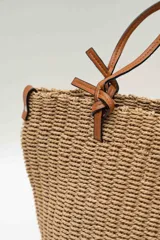 Bolso de mano tipo tote confeccionado en tejido texturizado efecto rafia en color beige, con asas dobles finas de cuero sintético marrón y detalles de nudos en los extremos.