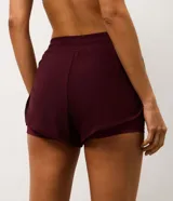 Bermuda deportiva femenina modelo runner, color bordó, con short interno incorporado. Presenta cintura elástica con cordón de ajuste y laterales con cordones ajustables. Confeccionada en tejido de tul con tecnología dry para mayor transpirabilidad.