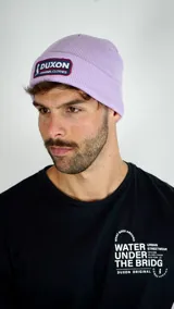 Gorro tipo beanie de tejido acanalado en color lila, con parche frontal rectangular de la marca Duxon.