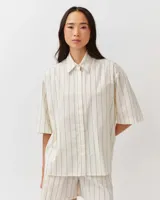 Camisa de corte recto y diseño depurado, color blanco con finas rayas verticales oscuras. Presenta cuello camisero clásico, cierre frontal de botones ocultos y mangas cortas de silueta relajada.