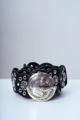 Cinto negro de cuero compuesto por piezas circulares unidas, con una gran hebilla metálica redonda plateada y detalles de tachas y argollas.