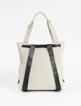 Mochila tipo shopper de tela color verde oliva, con asas de mano y bolsillo frontal con cierre de cordón. Presenta un detalle colgante de cordón naranja y verde neón en una de las asas.