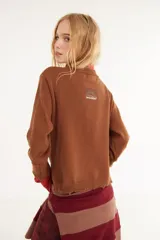 Sweater de punto color marrón con detalles en rojo en el cuello y los puños. Presenta un diseño de doble capa en el cuello y mangas largas con orificios para los pulgares, además de un pequeño bordado en el pecho y un acabado desflecado en el ruedo.