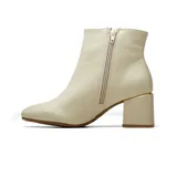 Bota de cuero color beige con taco cuadrado medio y detalle metálico dorado en el talón. Presenta un aplique triangular decorativo con strass en el lateral superior y cierre lateral.
