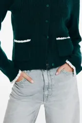 Saco corto de punto trenzado fino en color verde oscuro, con cuello redondo, cierre frontal de botones y bolsillos delanteros. Presenta detalles de ribetes festoneados en color crema en los bolsillos y en los puños.