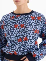 Camisa de viscosa con diseño estampado de corazones rojos sobre fondo azul con manchas. Presenta cuello redondo, puños y ruedo tejidos en ribb, con mangas largas abullonadas y hombros caídos.