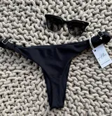 Bottom de bikini colaless negro, sin costuras, con textura de rayitas en la parte delantera y lycra con brillitos en la parte trasera. Breteles regulables en los costados.