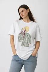 Remera de algodón de manga corta y cuello redondo, con estampado central que ilustra a una mujer de espaldas con pañuelo en el cabello y camisa a rayas.