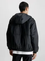 Campera cortavientos negra con capucha, confeccionada en poliéster reciclado. Cuenta con cierre de cremallera, puños y bajo elásticos, bolsillos laterales y logo de Calvin Klein en la manga.