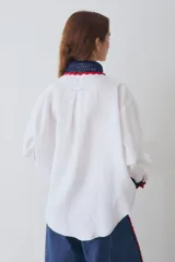 Camisa de lino blanca con cuello y puños tejidos a mano en crochet azul con borde rojo. Presenta cierre frontal con botones de madera, bolsillo frontal y costuras en contraste.