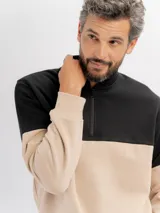Buzo de felpa con diseño color block, combinando negro en la parte superior y beige en la parte inferior. Presenta cuello alto con cierre frontal parcial y puños elásticos.