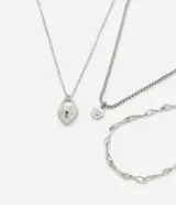 Set de tres collares en capas de metal plateado. Incluye una cadena de eslabones entrelazados, una cadena de eslabones cuadrados con un dije circular con strass, y una cadena fina con un dije de candado en forma de corazón.