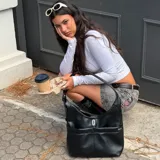 Cartera tipo hobo de color negro, confeccionada en material sintético con acabado liso. Presenta un diseño estructurado con dos bolsillos frontales funcionales y un detalle metálico central con el logo de la marca. Cuenta con una correa ajustable con hebilla y cierre principal.