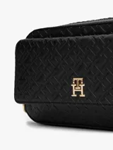 Cartera estilo camera bag negra, estructurada y compacta, con textura integral de monograma TH en relieve y placa metálica dorada con el logo TH en el frente. Posee cierre superior y correa ajustable para llevar al hombro o cruzada.
