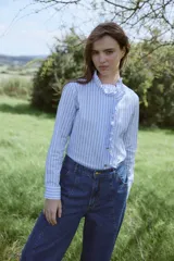 Camisa de algodón con estampado de rayas verticales en color bordó y blanco. Presenta un diseño de manga larga con puños abotonados, cuello camisero y un detalle de volado lateral que recorre el frente desde el cuello hasta el ruedo, acompañando la línea de botones.
