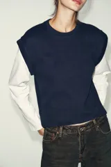 Prenda de manga larga blanca con un chaleco de tejido de punto azul marino superpuesto, creando un efecto de dos piezas. El chaleco es de cuello redondo y corte recto.