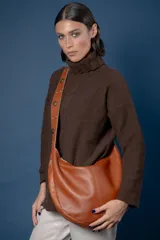 Bolso tipo hobo de cuero color suela, con correa larga ajustable decorada con tachas metálicas. Presenta un diseño curvo y minimalista con cierre superior.