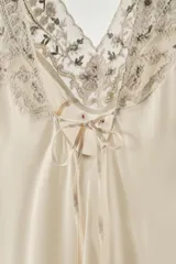 Vestido mini tipo lencero de satén color crema, con escote en pico y tirantes anchos. Presenta bordados florales detallados con aplicación de abalorios, flores y lazos en relieve a contraste en el escote y el ruedo. Cierre en la espalda con lazada.