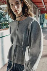Jersey de punto color gris, con cuello redondo, mangas largas abullonadas con puños elásticos y cintura ajustable mediante cordón. Presenta una costura vertical marcada en la parte frontal.