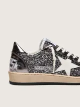 Zapatillas Golden Goose modelo Ball Star de estilo retro, con empeine y lengüeta cubiertos de glitter gris oscuro y detalles en cuero laminado. Presentan la clásica estrella lateral en cuero blanco, puntera reforzada y cordones de algodón crudo, con suela de goma desgastada.