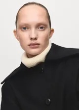 Chaqueta negra de corte corto y holgado, confeccionada con mezcla de lana. Cuenta con cuello ancho y solapa de piel sintética en tono beige claro, cierre frontal con botones oscuros y mangas largas amplias.