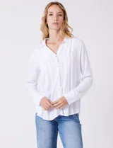 Camisa blanca de rayón con cuello solapa, escote en V y cierre frontal con botones. Presenta plisados verticales en la parte delantera y un canesú con tablón central en la espalda. El bajo es redondeado y ligeramente más largo en la espalda.