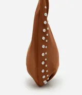 Cartera tipo baguette confeccionada en gamuza sintética color marrón. Presenta un diseño curvo con aplicaciones de tachas metálicas plateadas en los laterales, asa corta integrada y cierre mediante botón a presión.