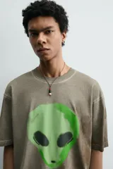 Camiseta de corte relajado con cuello redondo y manga corta. Presenta un efecto de lavado desgastado en tonos marrones y un estampado frontal de una cabeza de alienígena en color verde vibrante.