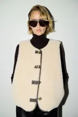 Chaleco de efecto peludito color beige, con cuello redondo, cierre frontal de botones y ribetes en contraste marrón oscuro. Incluye bolsillos laterales discretos y forro interior.