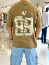 Remera regular color off-white, con diseño gráfico en la espalda que incluye el número 99 en gran tamaño y detalles en negro. Cuenta con ribetes negros en las costuras de los hombros y mangas.