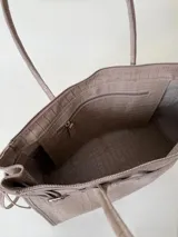 Bolso grande de cuero marrón con textura tipo cocodrilo, estructura rígida y asas para llevar al hombro. Tiene una tira con tachas y hebillas en cada extremo, también lleva lazos en los costados.