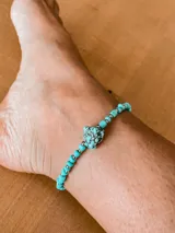 Tobillera elástica con cuentas de color turquesa y cuentas plateadas. Incluye una cuenta central de mayor tamaño con diseño jaspeado en negro.