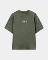 Remera oversize gris oscuro con corte holgado, mangas cortas y estampa trasera en blanco y negro con forma de estampilla que muestra a una persona con un gato y el texto 'Kaboa Industries'.