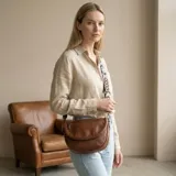 Cartera bandolera de eco-cuero marrón, de diseño redondeado con solapa frontal y bolsillo con cierre. Incluye correa ajustable y desmontable con patrón geométrico.
