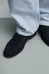 Mocasín de vestir para hombre, fabricado en piel con acabado en serraje de color azul marino. Presenta un diseño clásico con detalle de antifaz en el empeine, horma redonda, costuras visibles en el contorno y una suela sutil con ligero tacón.