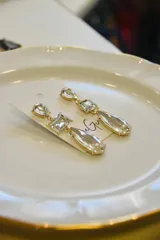 Par de aros colgantes dorados con tres piedras facetadas transparentes de diferentes formas: gota, rectángulo y lágrima.